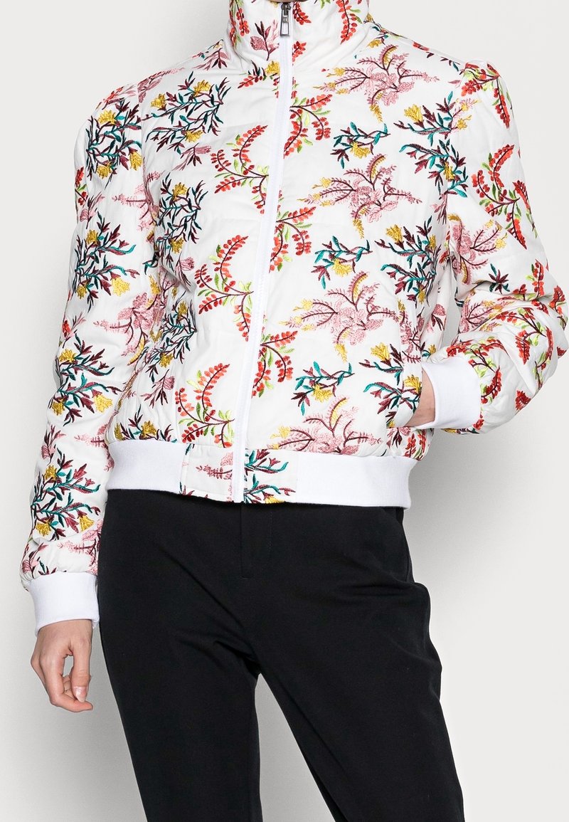Veste blanche zippée avec broderie florale colorée, portée avec un pantalon noir, main dans la poche, debout devant un arrière-plan uni.