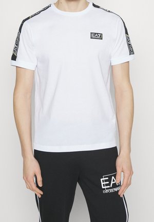 Mężczyzna w białym T-shircie EA7 Emporio Armani z czarnymi markowymi paskami na ramionach i czarnych spodniach z białym logo i obramowaniem.