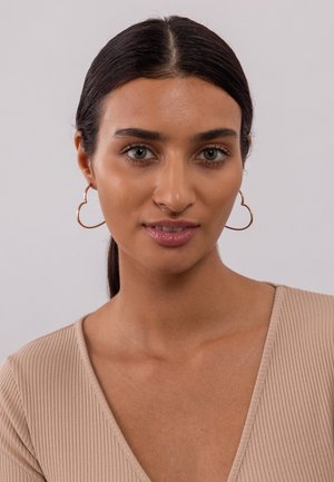 Gouden hartvormige hoepeloorbellen met een gladde afwerking, gedragen door een model met steil donker haar en een beige geribbelde top.