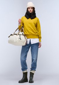 Sudadera amarilla, bufanda de plumas negra, camisa blanca, jeans azules, botas de goma verdes, bolso grande color crema con asas beige y acentos negros.