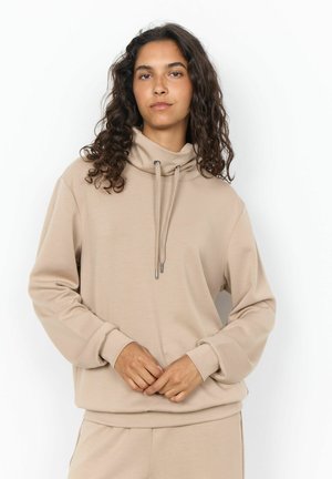 Giovane donna con capelli ricci che indossa una felpa beige con collo alto e pantaloni coordinati, in piedi con le mani leggermente intrecciate.