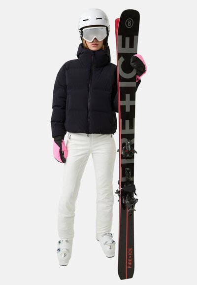 Zwarte pufferjas met een hoge kraag, witte strakke skibroek, roze handschoenen, witte skihelm, spiegelende skibril en zwarte ski's met rode accenten.