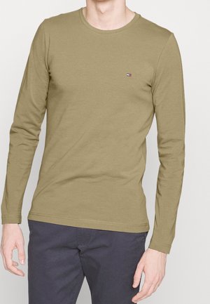 Mann trägt ein olivgrünes Langarm-Shirt mit Rundhalsausschnitt und kleinem Logo auf der Brust, kombiniert mit dunkelgrauen Hosen, steht vor hellem Hintergrund.