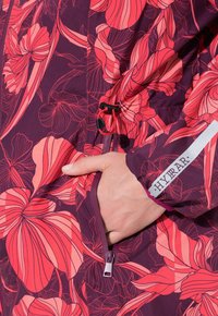 Ruka v kapse fialového květinového saka s růžovými a červenými abstraktními květinovými vzory a černou šňůrkou s táhlem poblíž kapsy.