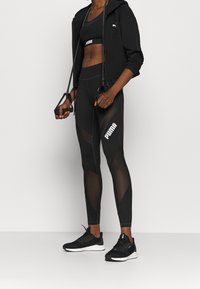 Completo sportivo nero composto da un hoodie corto, un reggiseno sportivo e leggings con dettagli in rete, abbinato a scarpe da corsa nere.