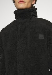 Černá fleecová bunda s vysokým límcem, s texturovaným povrchem a malou obdélníkovou nášivkou na hrudi.