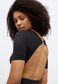 Μαύρο κορμάκι με κοντά μανίκια και βαθύ άνοιγμα στην πλάτη, με χρυσή λεπτομέρεια στον λαιμό. Λείο ύφασμα, εφαρμοστό σχέδιο.