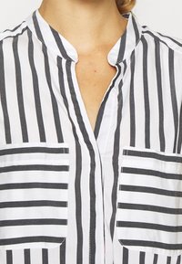 Camicia button-up in tessuto leggero, caratterizzata da strisce verticali bianche e nere, un collo senza colletto e due taschini sul petto.