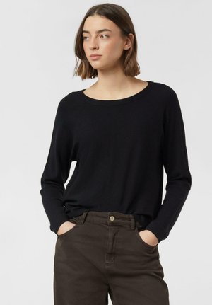 Młoda kobieta o brązowych włosach do ramion, ubrana w czarną bluzkę z długim rękawem i brązowe spodnie, ręce w kieszeniach, patrzy w lewo.