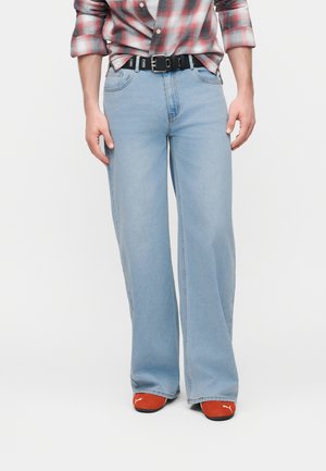 Άνδρας που φοράει ανοιχτό μπλε τζιν παντελόνι με φαρδιά μπατζάκια, μαύρη ζώνη, κόκκινα αθλητικά παπούτσια Puma και κόκκινο-λευκό πουκάμισο καρό με ανασηκωμένα μανίκια.