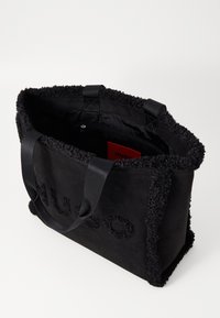 Sac fourre-tout noir à texture douce, avec une bordure duveteuse et un logo embossé. Double anses en tissu et poche intérieure zippée.