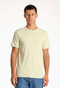 T-shirt di cotone giallo chiaro con maniche corte, scollatura rotonda e testo del logo frontale in bianco. Abbinata a jeans blu.