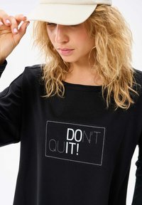 Czarna koszula z długim rękawem z białą prostokątną grafiką z napisem "NIE PODDAWAJ SIĘ!" w pogrubionej czcionce. Miękka tkanina. Casualowy design.