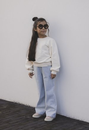 Witte sweatshirt met pofmouwen, blauwe verticaal gestreepte wijde broek, en grote zwarte zonnebril in de vorm van een bloem. Beige instap schoenen.