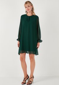 Zelena chiffon haljina s dugim prozirnim rukavima, okruglim dekolteom i naborima. Labav kroj iznad koljena. Crne sandale na visoku petu s trakama.