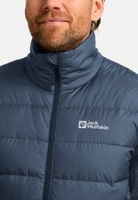 Blauer Pufferjacke mit hohem Kragen, vertikaler Steppoptik, durchgehendem Reißverschluss und einem weißen Logo auf der linken Brust.
