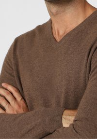 Brauner Wollmischpulli mit V-Ausschnitt, gerippten Bündchen und weicher Textur. Nahaufnahme, die auf die Brust und die verschränkten Arme im Vordergrund fokussiert.