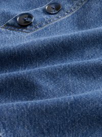 Denimtyg i blått med en mjuk textur. Har två mörka knappar och synlig sömnad, vilket visar en slät och något rynkad yta.