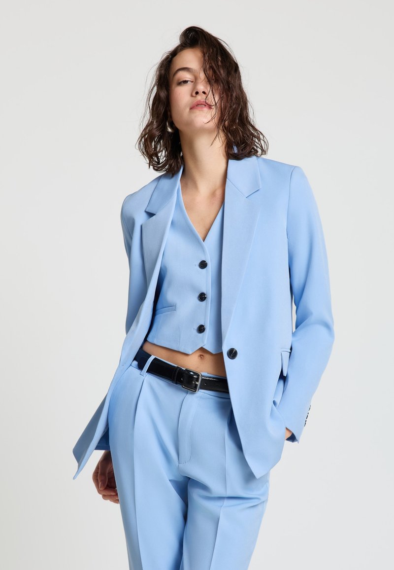 Jeune femme aux cheveux mouillés portant un costume bleu clair composé d'un gilet court, d'un blazer et d'un pantalon, posant avec une main dans sa poche.