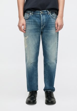 Jean en denim bleu clair à coupe décontractée, présentant des zones usées, un léger délavage et un design cinq poches. Porté avec des chaussures noires.