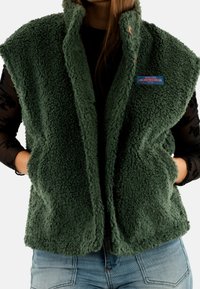 Gilet di pile verde con colletto alto, tasche laterali e un'etichetta blu. La texture è morbida e soffice, con un design casual senza maniche.