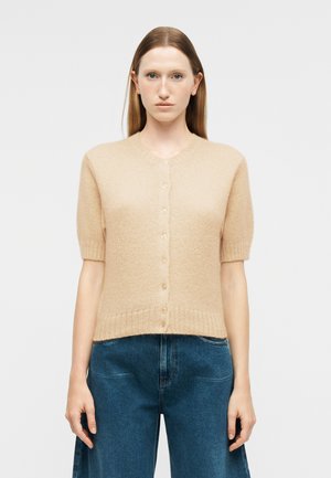 Donna di fronte che indossa un cardigan beige a maniche corte con bottoni e jeans blu a vita alta, su uno sfondo semplice.