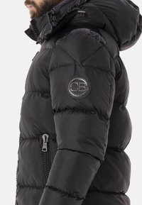 Schwarze Pufferjacke mit gestepptem Design, ausgestattet mit einem Reißverschluss vorne und einer Kapuze. Silbernes "CB"-Logo auf dem linken Ärmel, glatte Stoffstruktur.