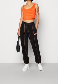 Vrouw in een oranje mouwloos crop top, zwarte joggers met oranje stiksels, witte platform sneakers, en een kleine zwarte tas.