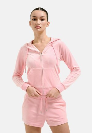 Vrouw draagt een lichtroze hoodie met ritssluiting en bijpassende korte broek, staand met handen bij de tailleband tegen een effen achtergrond.