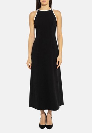 Donna che indossa un abito midi nero senza maniche con bordino bianco sulla scollatura e tacchi neri, in piedi davanti a uno sfondo semplice.