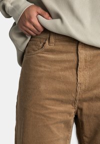 Ręka podciągająca beżowy sweter o splotach wafelowych wsunięty w jasnobrązowe spodnie z kordurowej tkaniny.