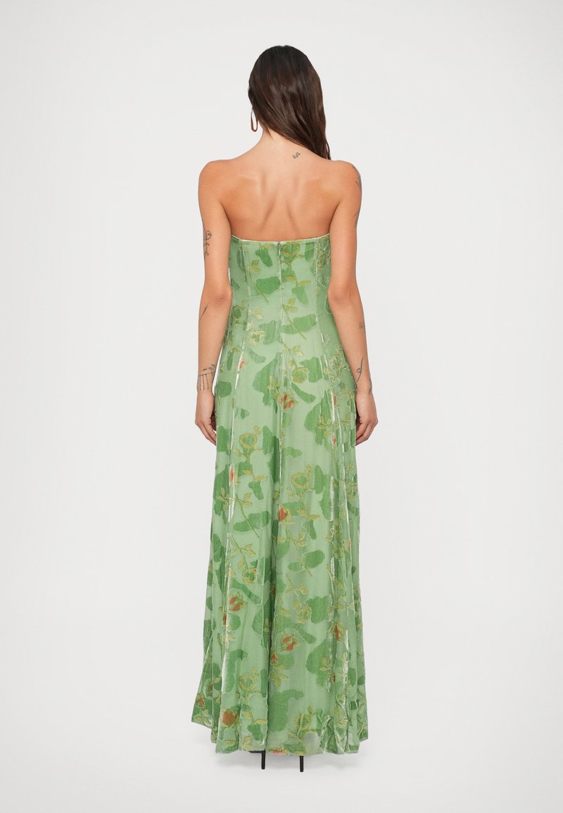 Woman with long dark hair and tattoos wears strapless green floral maxi dress, standing with back to camera against white background.
