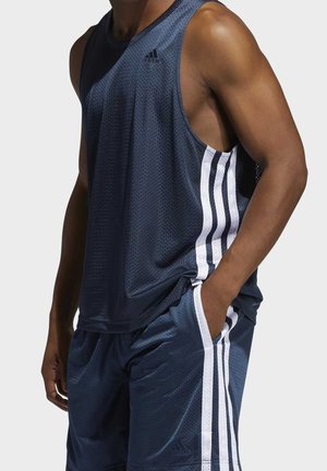 Mężczyzna w granatowym bezrękawniku sportowym Adidas i pasujących szortach z białymi paskami, ręka w kieszeni.
