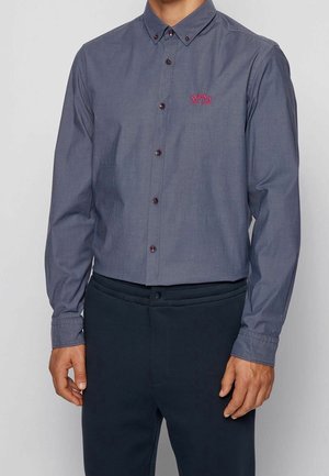 Mężczyzna w szarej koszuli na guziki z długim rękawem, z czerwonymi guzikami i czerwonym logo „BOSS” na piersi, w zestawie z ciemnymi spodniami.