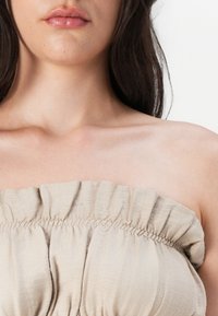 Beige skulderløs topp med samlet volangtekstur. Mykt, lett stoff komplementerer en jevn halslinje og kragebein.