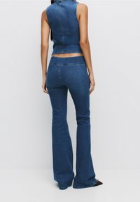 La tenue en denim se compose d'un haut sans manches et de jeans évasés d'une couleur bleu moyen. Le tissu a une texture lisse avec des détails de couture visibles.