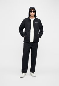 Άνδρας με μαύρο μπουφάν με κουκούλα, μαύρο παντελόνι, λευκό πουκάμισο, λευκά αθλητικά παπούτσια και μαύρα γυαλιά ηλίου, στέκεται με τα χέρια στις τσέπες.