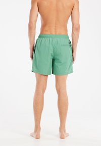Shorts de bain verts en tissu léger, dotés d'une taille élastique, de poches latérales et d'une poche au dos. Longueur courte avec une texture lisse.