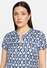 Blouse à manches courtes présentant un motif ondulé multicolore en tons de bleu, crème et pêche ; le tissu semble lisse avec un col à revers.
