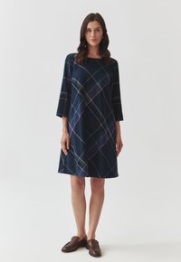Robe marine à manches trois quarts, ornée d'un motif à carreaux en vert, rose et blanc. Tissu doux et forme évasée. Chaussures brunes à enfiler.