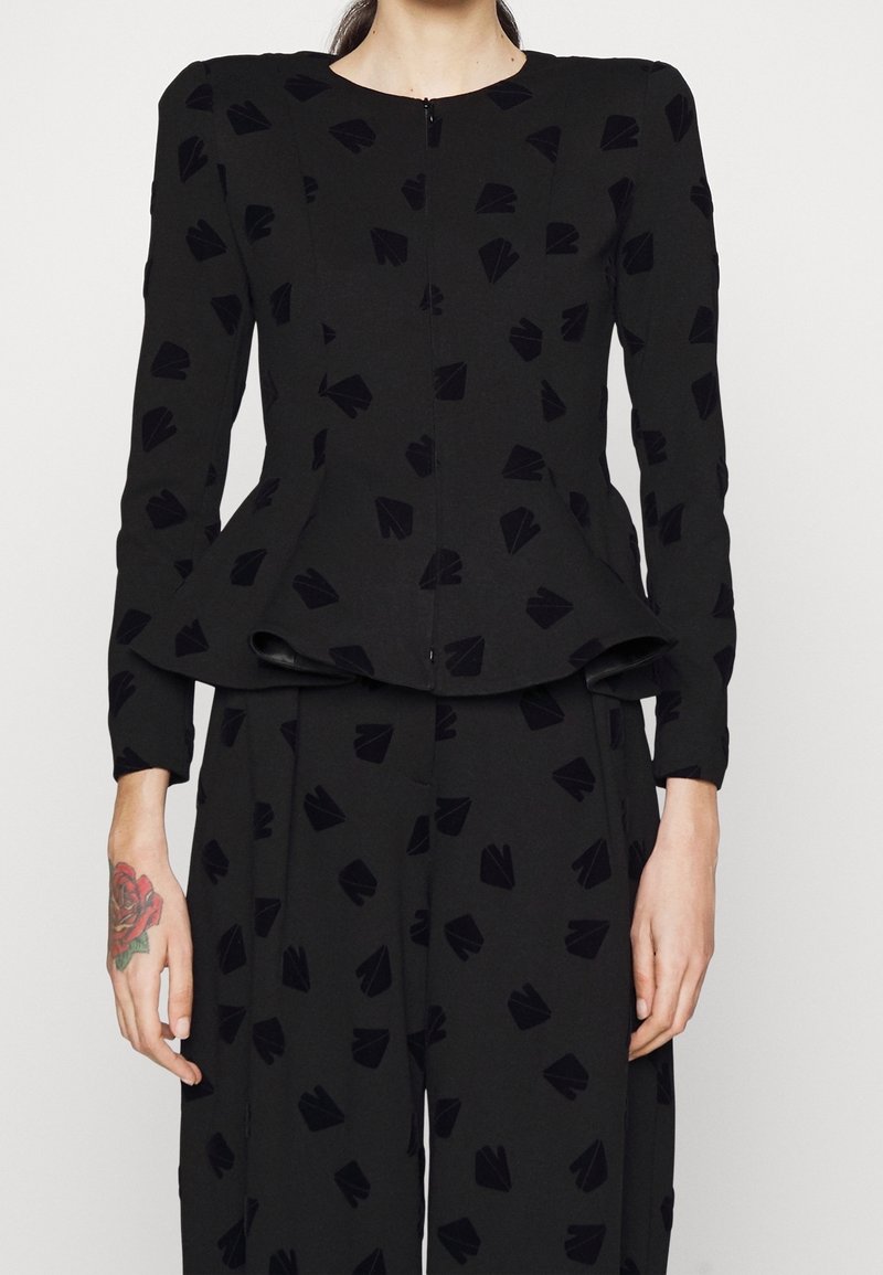 Černý peplum top s dlouhými rukávy, texturovaná látka s vzorem drobných tmavých tvarů a předním zapínáním na zip.