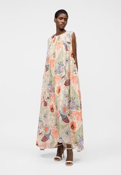 Γυναίκα που φοράει αμάνικο, μακρύ μέχρι το πάτωμα φόρεμα με άνθη σε παστέλ αποχρώσεις πορτοκαλί, μπλε, πράσινο και κρεμ, συνδυασμένο με μπεζ πέδιλα με ψηλό τακούνι.