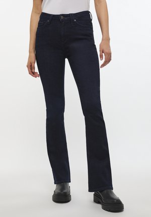 Person iført mørkeblå slim bootcut jeans, sorte chunky sko og en hvid top, stående mod en hvid baggrund.