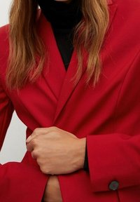 Henkilö, joka pitää kirkkaita punaisia bleisereitä mustan päälliset yli ja sulkee bleiserin yhdellä kädellä vyötäröllä.