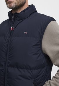 Marineblå puffervest med høy krage, glidelåslukking og kontrasterende logo-lapp. Har teksturert stoff og sidelommer for praktisk bruk.