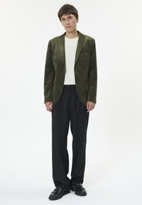 Olivově zelený sametový blazer s klopami a kapsou, kombinovaný s bílým tričkem a černými kalhotami, doplněný černými botami.