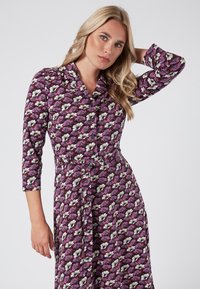 Vestido morado con un patrón floral que presenta flores blancas y rosas, mangas tres cuartos, un cuello y una cintura Ceñida. Textura de tela suave.
