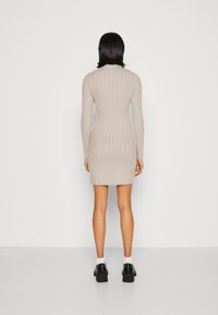 Abito beige a coste con maniche lunghe, silhouette aderente, lunghezza al ginocchio e sottile texture verticale. Indossato con scarpe nere con tacco largo.