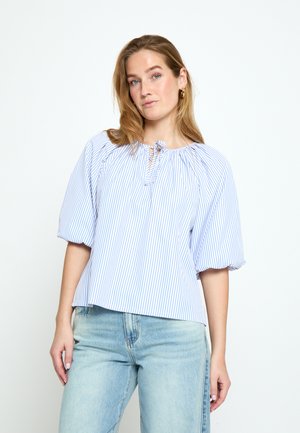 Mladá žena oblečená v modro-bielej pruhovanej blúzke s nafúknutými rukávmi a svetlomodrých džínsoch, stojaca pred jednoliatym bielym pozadím.