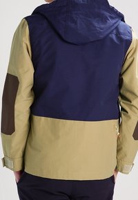 Kurtka z blokami kolorów, z granatową górną częścią, khaki dolną częścią oraz brązowymi łatami na łokciach. Z regulowaną kapturem i mankietami.